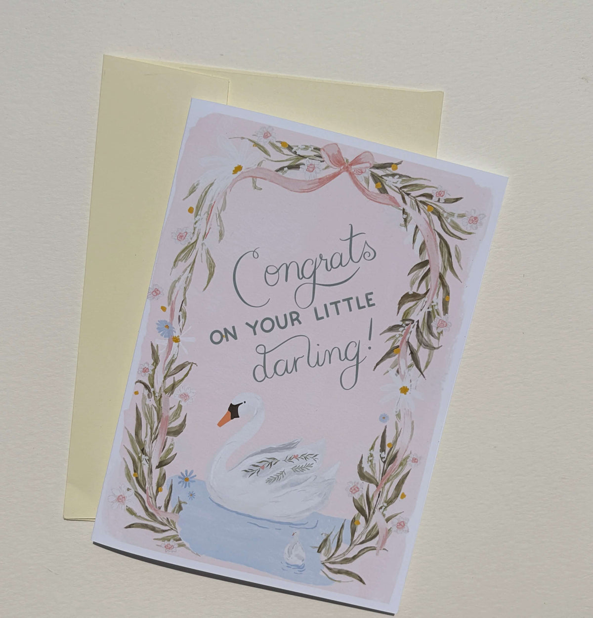 A congratulations card for a baby swan, featuring a watercolor illustration of a white swan in a blue pond, surrounded by a floral wreath and the text 'Congrats on your little darling!'. The card is shown with a pale yellow envelope on a light beige background.
