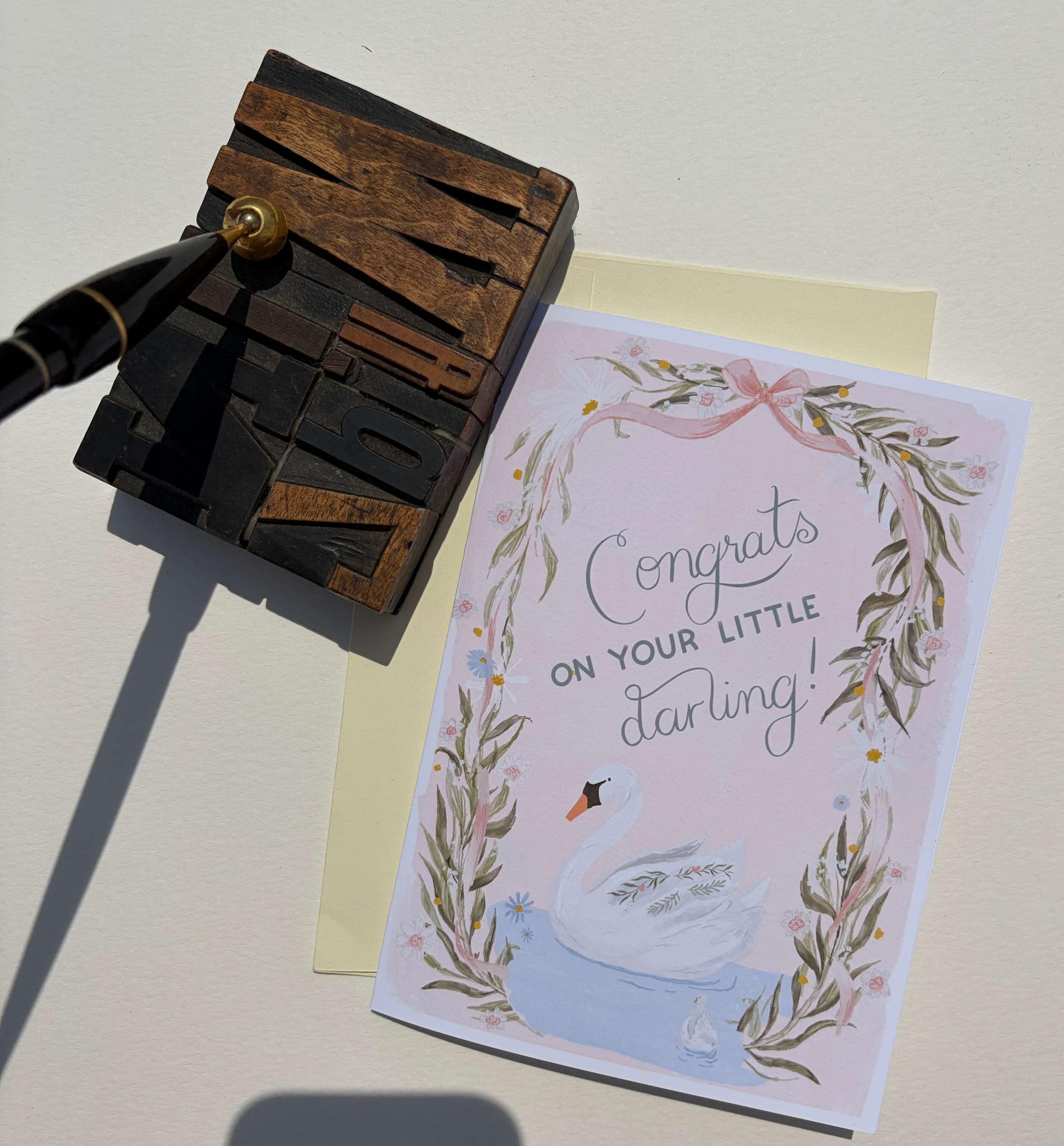 A congratulations card for a baby swan, featuring a watercolor swan and leaf wreath on a pale pink background with the text 'Congrats on your little darling!'. The card is styled on a cream surface next to a wooden letterpress block and a black fountain pen.