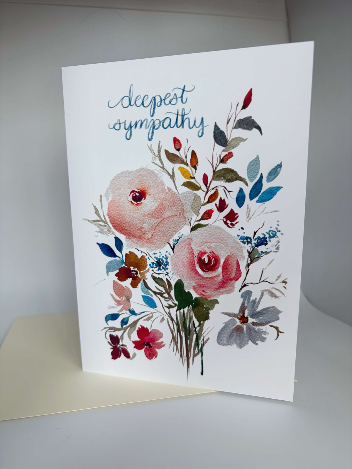 A white sympathy greeting card standing upright, printed with the words 'deepest sympathy' in blue script above a watercolor floral arrangement in pink, blue, red, green, and gray tones.