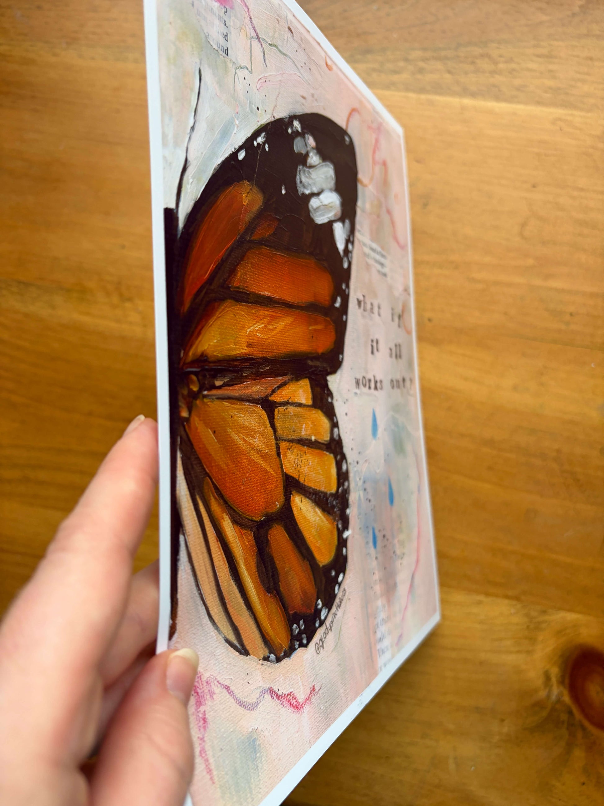 A hand holding a vertical art print featuring a painted monarch butterfly wing in orange, black, and cream tones, photographed over a wooden tabletop.
