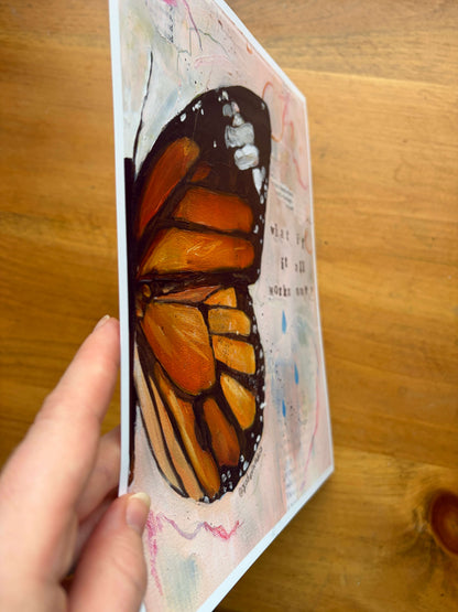 A hand holding a vertical art print featuring a painted monarch butterfly wing in orange, black, and cream tones, photographed over a wooden tabletop.