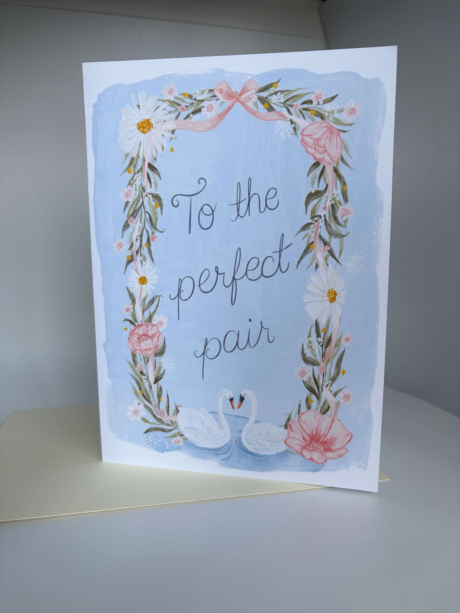 A 5x7 greeting card with a watercolor-style floral border and two swans, reading 'To the perfect pair,' displayed upright on a light surface against a neutral gray background.