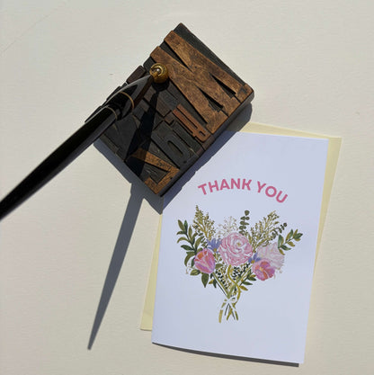 A white thank you card with pink text and a floral bouquet illustration, placed on a cream envelope next to a wooden letterpress block with a black ink pen resting on top, all on a light beige background.