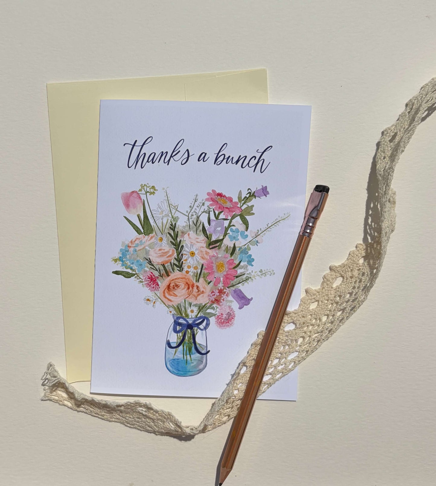 A greeting card that reads 'thanks a bunch' with a watercolor bouquet in a blue vase, shown with a cream envelope, a wooden pencil, and a piece of lace on a light beige background.