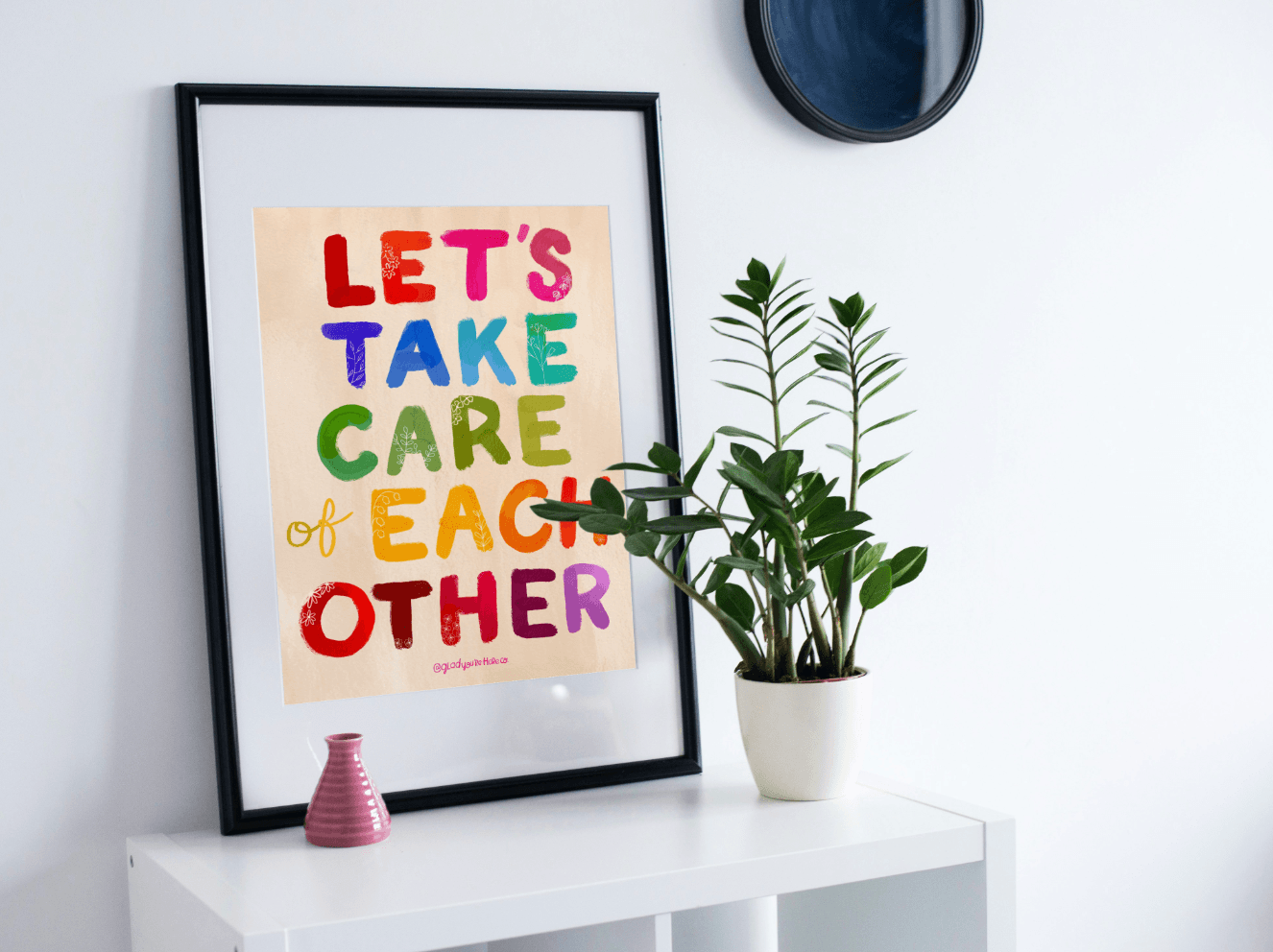 Framed colorful poster with 'Let's Take Care of Each Other' text on a white wall with a plant and vase.