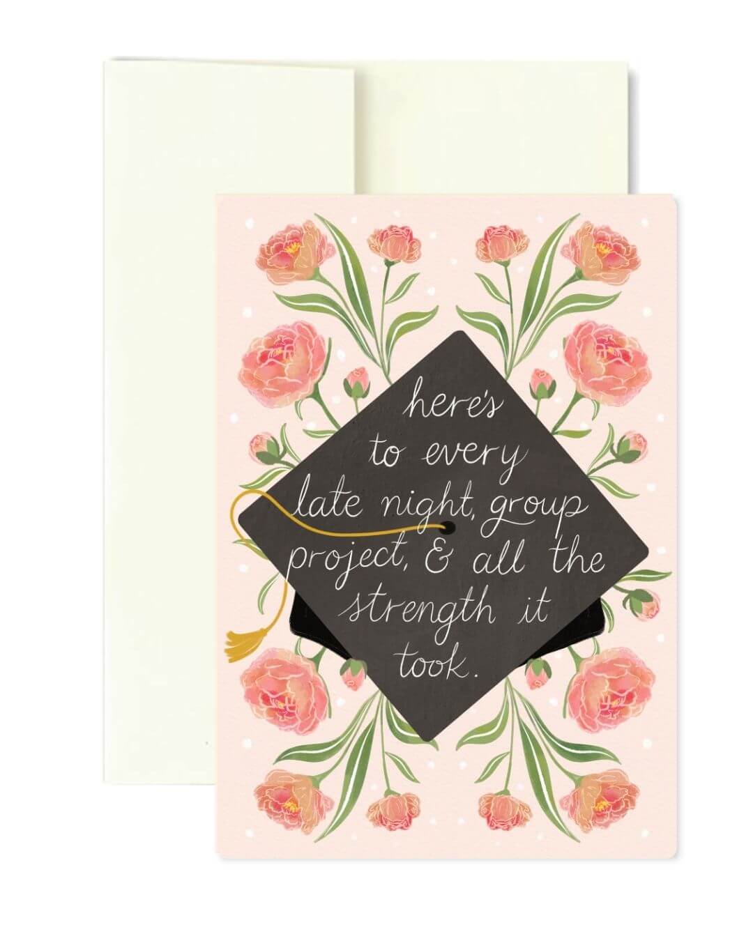 A graduation-themed greeting card with a pink floral border and a black mortarboard cap graphic in the center, overlaid with the phrase, 'here's to every late night, group project, & all the strength it took.' A cream envelope is visible behind the card on a white background.