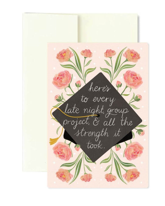 A graduation-themed greeting card with a pink floral border and a black mortarboard cap graphic in the center, overlaid with the phrase, 'here's to every late night, group project, & all the strength it took.' A cream envelope is visible behind the card on a white background.