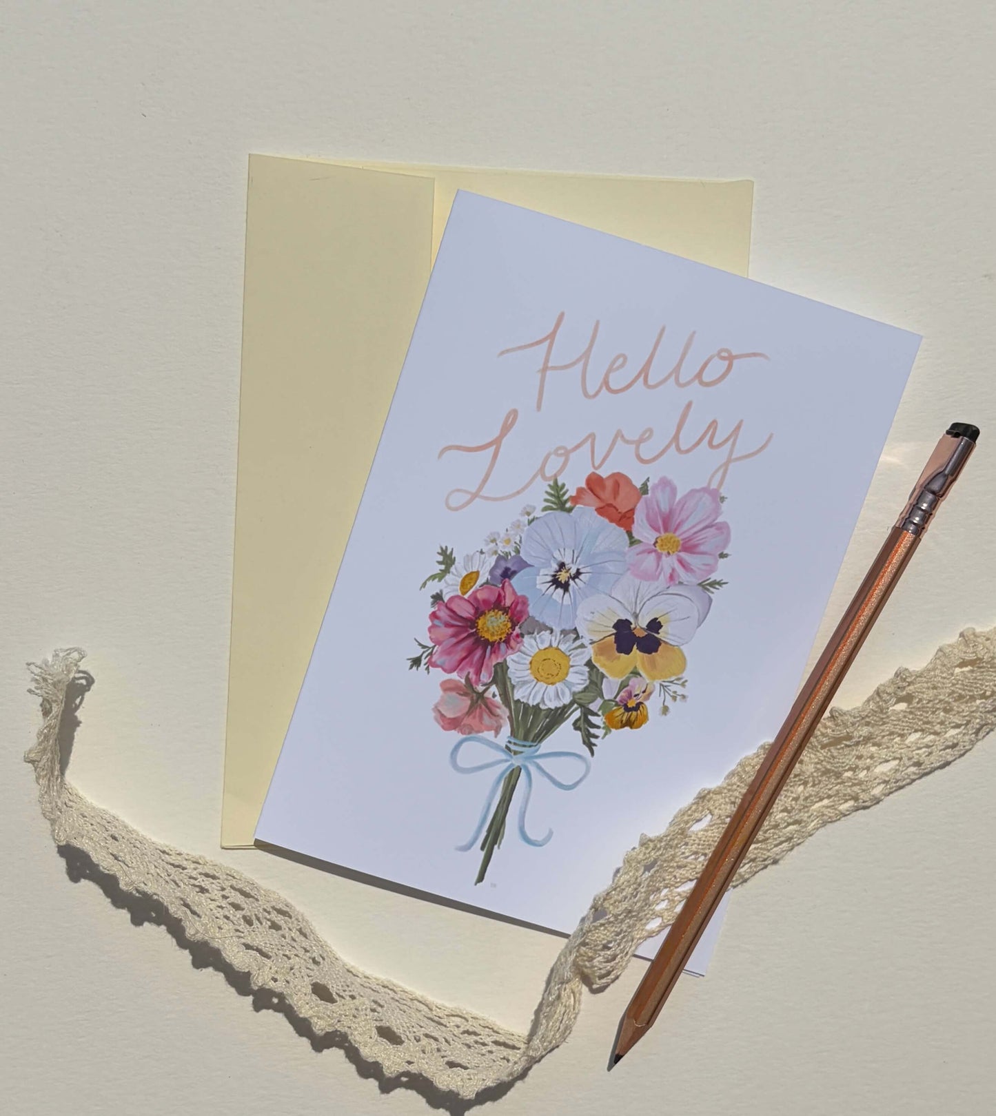 A square greeting card with a pastel floral bouquet illustration and the words 'Hello Lovely' in rose-gold script, shown with a cream envelope, a wooden pencil, and a piece of lace on a warm off-white background.