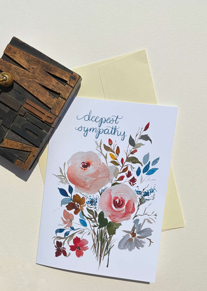 A sympathy greeting card with a watercolor floral design and the words 'deepest sympathy' in blue script, shown with a pale yellow envelope and a wooden letterpress block on a light beige background.