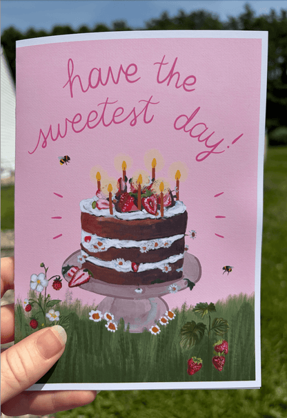 A hand holds a pink birthday card outdoors. The card features an illustration of a two-tier chocolate cake with candles on a stand, surrounded by grass, flowers, strawberries, and bees, with the text 'have the sweetest day!' in pink script at the top.