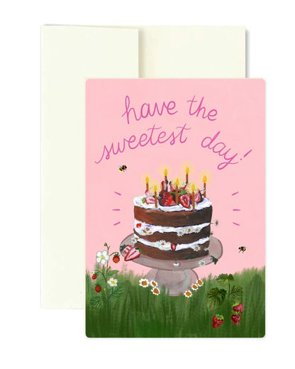 A birthday greeting card with a pink background and the text 'have the sweetest day!' above an illustration of a chocolate cake on a stand in a grassy field. A cream envelope is placed behind the card.