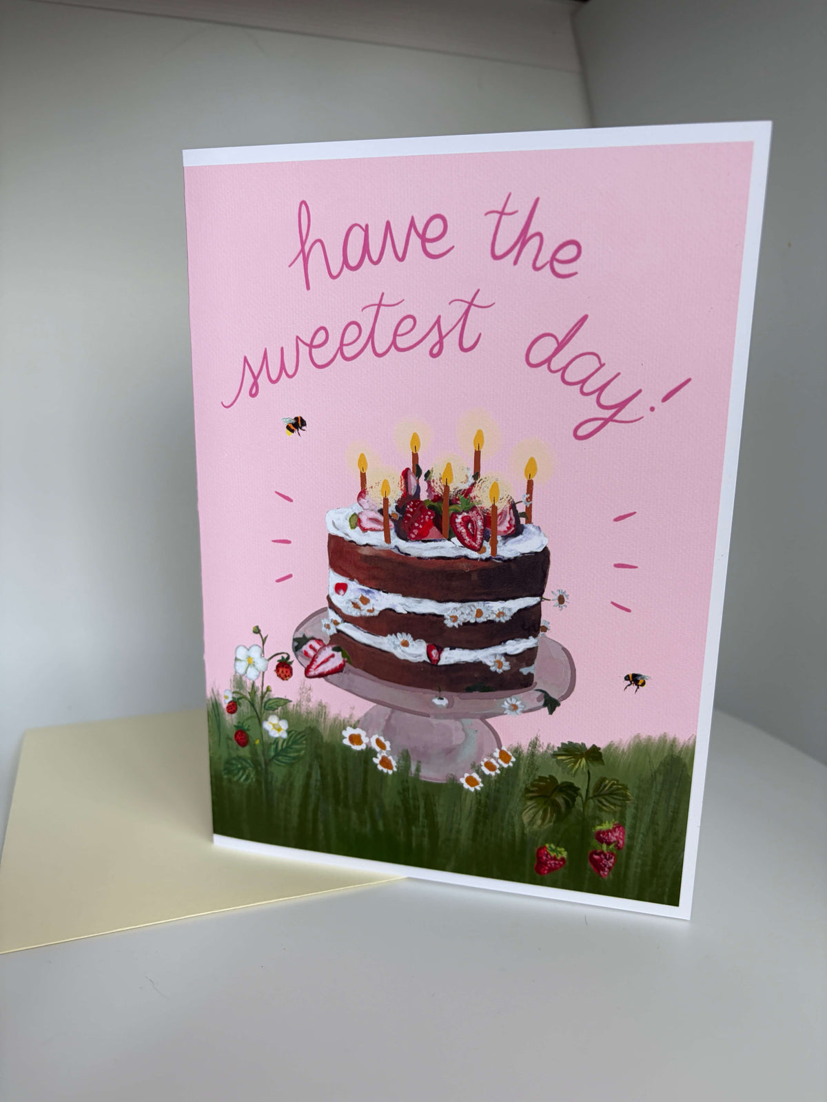 A pink birthday card with the handwritten text 'have the sweetest day!' above an illustration of a chocolate cake with candles, set on a white surface with a cream envelope behind it.