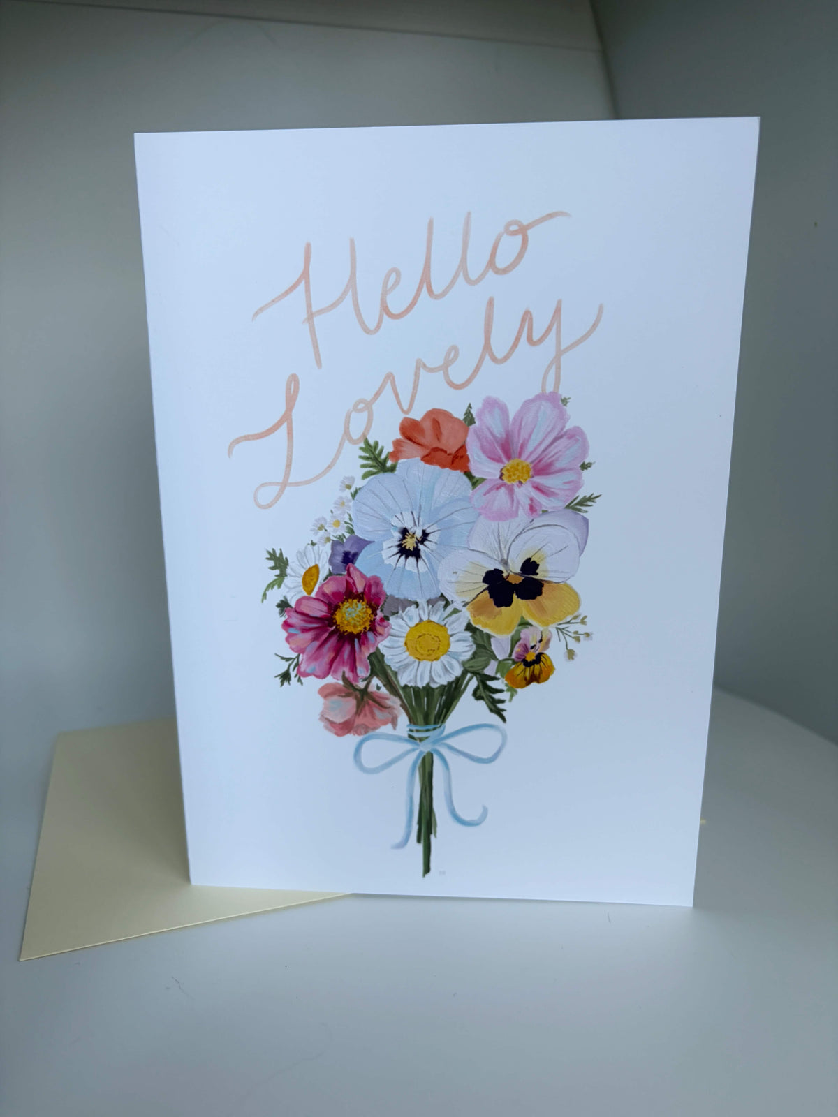 A white greeting card with the words 'Hello Lovely' in a rose-gold metallic script font, above a colorful watercolor bouquet of flowers including pink, blue, yellow, and orange blossoms tied with a light blue ribbon. A cream-colored envelope is partially visible underneath the card on a light gray surface.