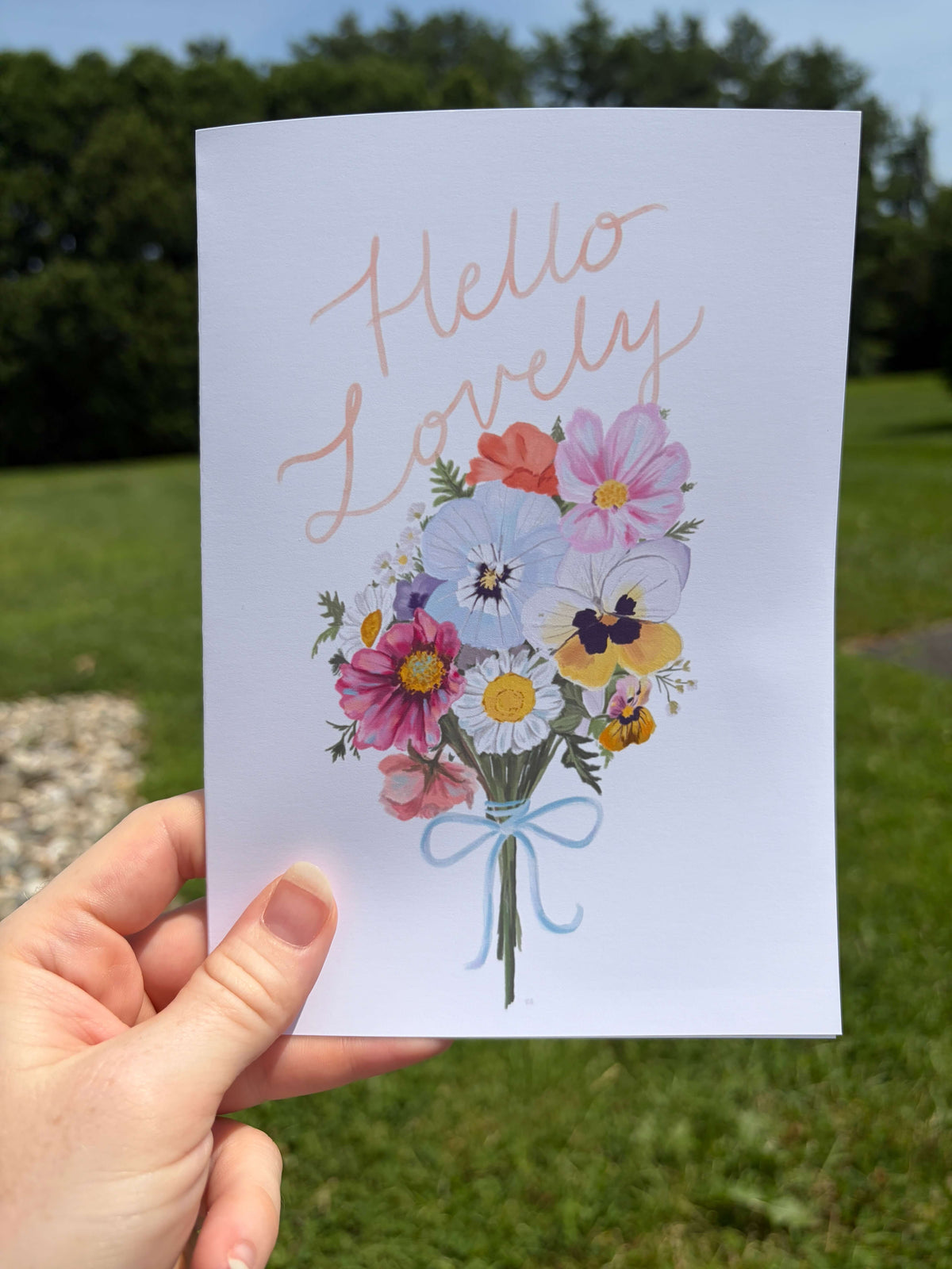 A hand holds a white greeting card outdoors with a blurred green lawn and trees in the background. The card features the words 'Hello Lovely' in a dusty rose script above a colorful bouquet of wildflowers tied with a light blue ribbon.