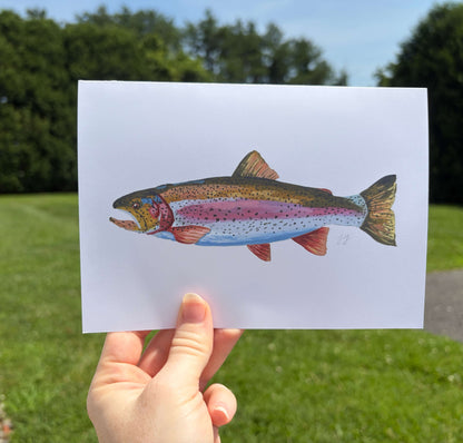 A hand holds a white art print featuring a detailed illustration of a rainbow trout against a blurred outdoor background of green grass and trees.