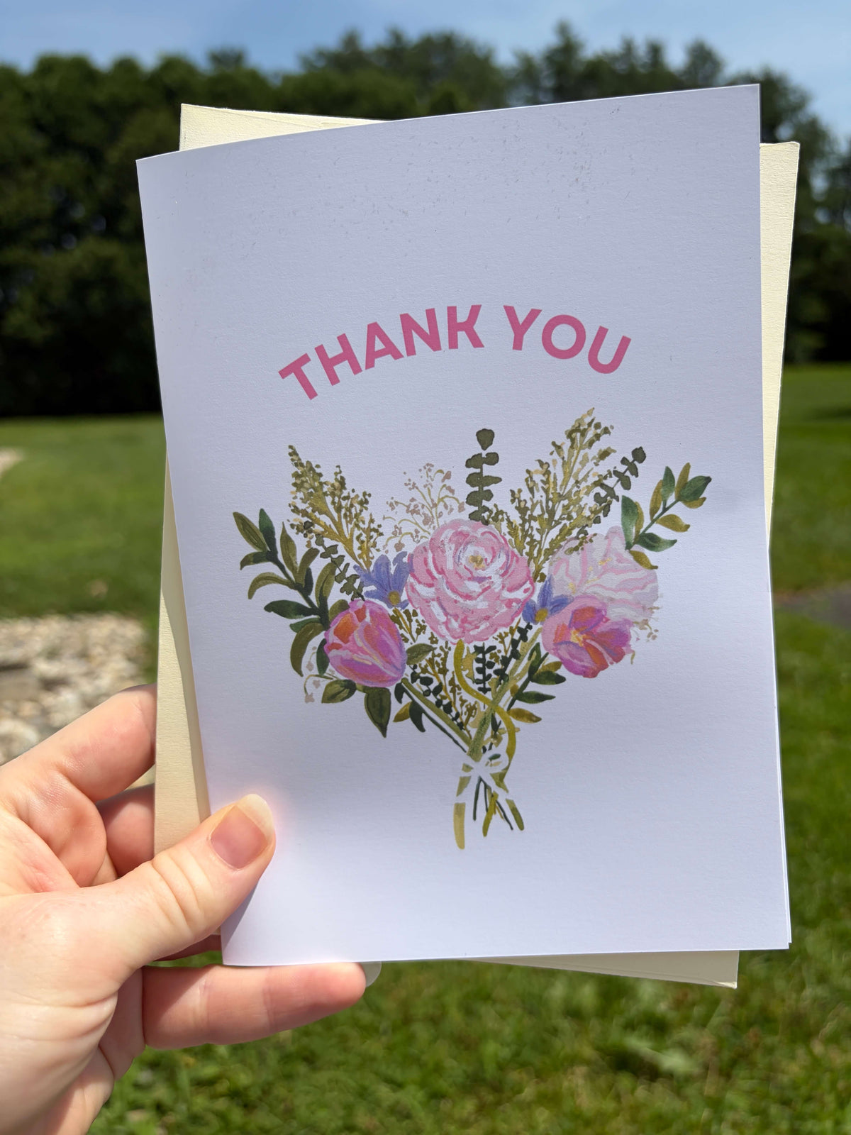 A hand holds a white thank-you card outdoors. The card has pink 'THANK YOU' text above a watercolor-style bouquet of pink and purple flowers with green leaves.
