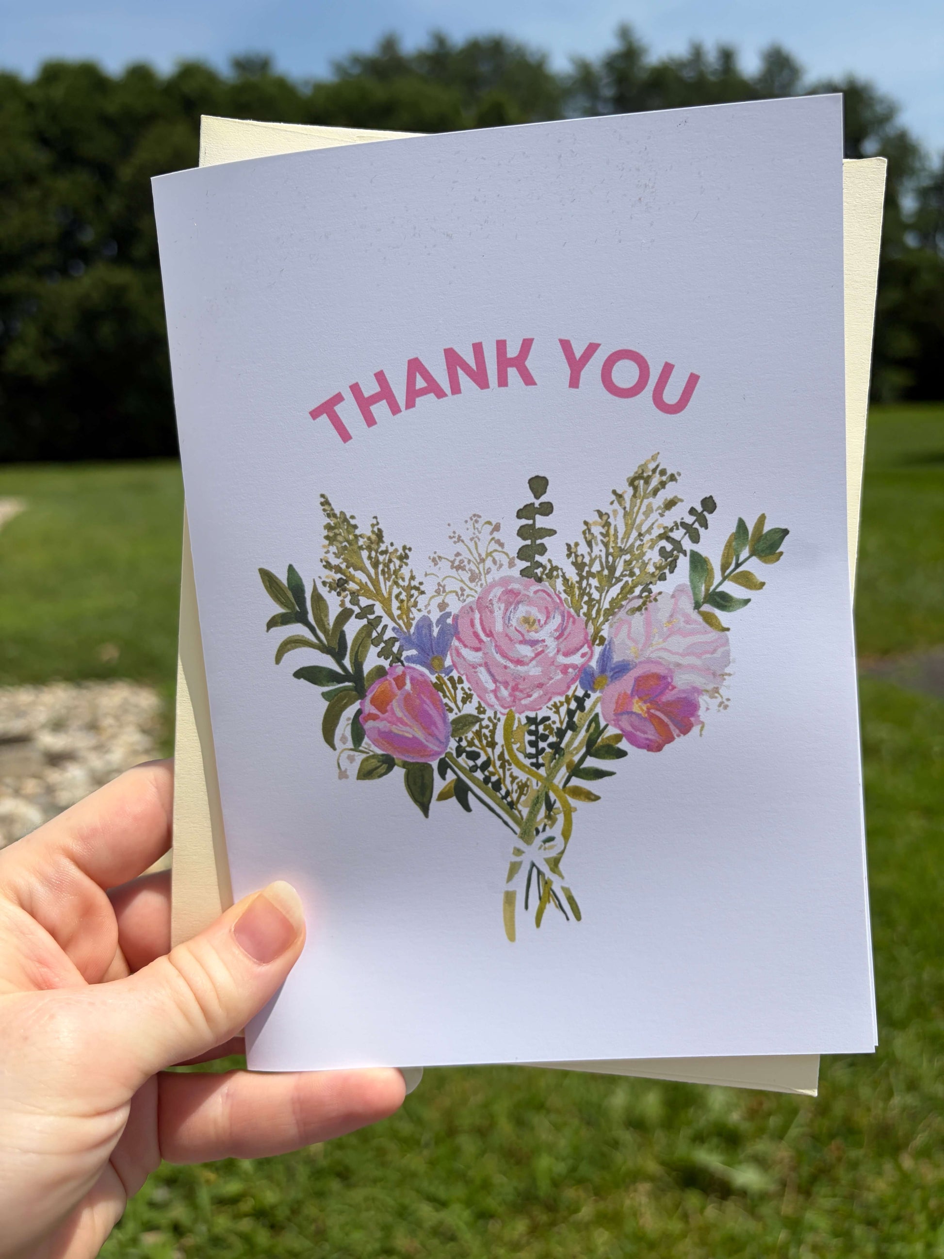 A hand holds a white thank-you card outdoors. The card has pink 'THANK YOU' text above a watercolor-style bouquet of pink and purple flowers with green leaves.