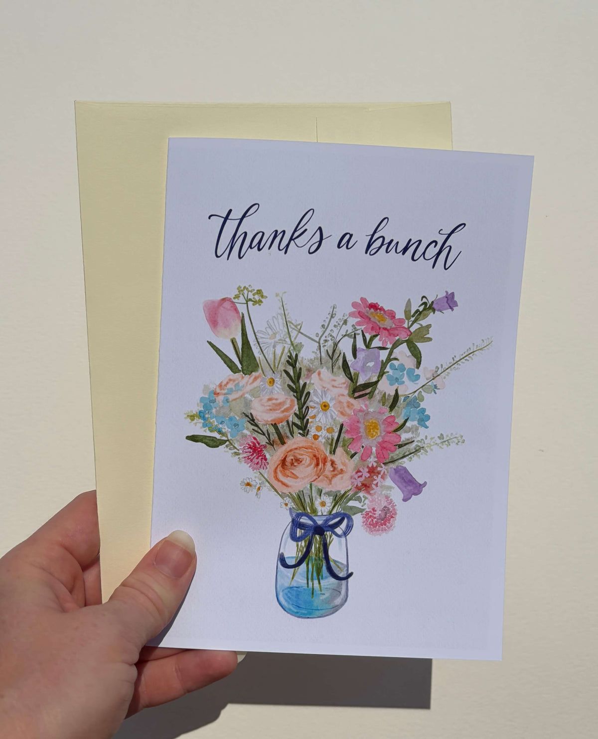 A hand holding a white greeting card that says 'thanks a bunch' above a watercolor-style bouquet of pink, peach, blue, and purple flowers in a glass jar, with a pale yellow envelope behind it.