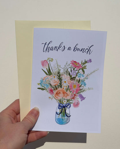 A hand holding a white greeting card that says 'thanks a bunch' above a watercolor-style bouquet of pink, peach, blue, and purple flowers in a glass jar, with a pale yellow envelope behind it.