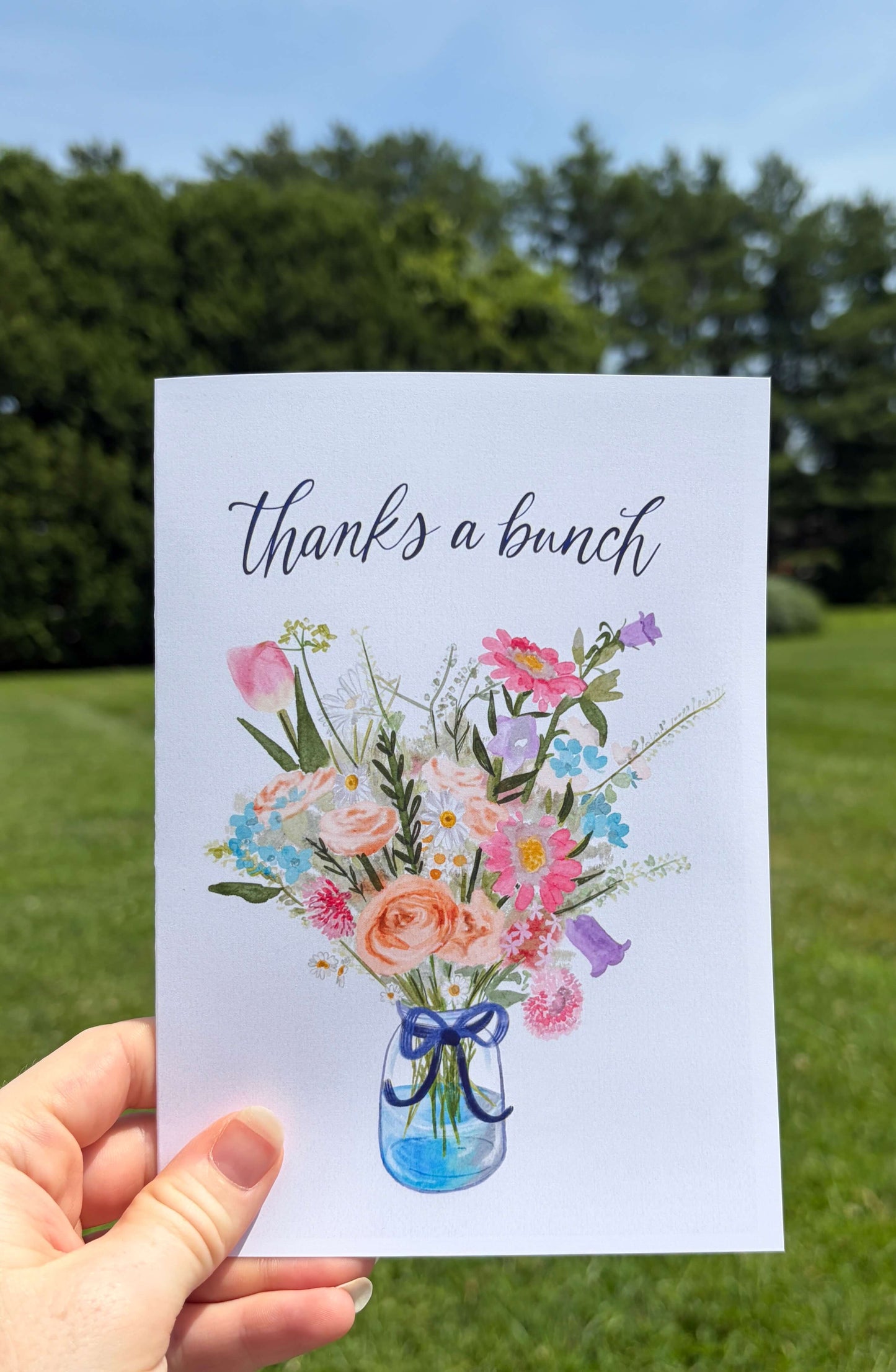 A hand holds a white greeting card outdoors against a blurred green lawn and blue sky. The card reads 'thanks a bunch' above a watercolor-style bouquet of pink, peach, blue, purple, and green flowers in a blue glass vase.