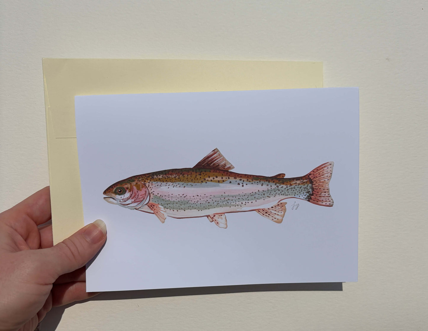 A hand holds a white art print or postcard featuring a detailed illustration of a rainbow trout, with a pale yellow backing card behind it against a light beige wall.