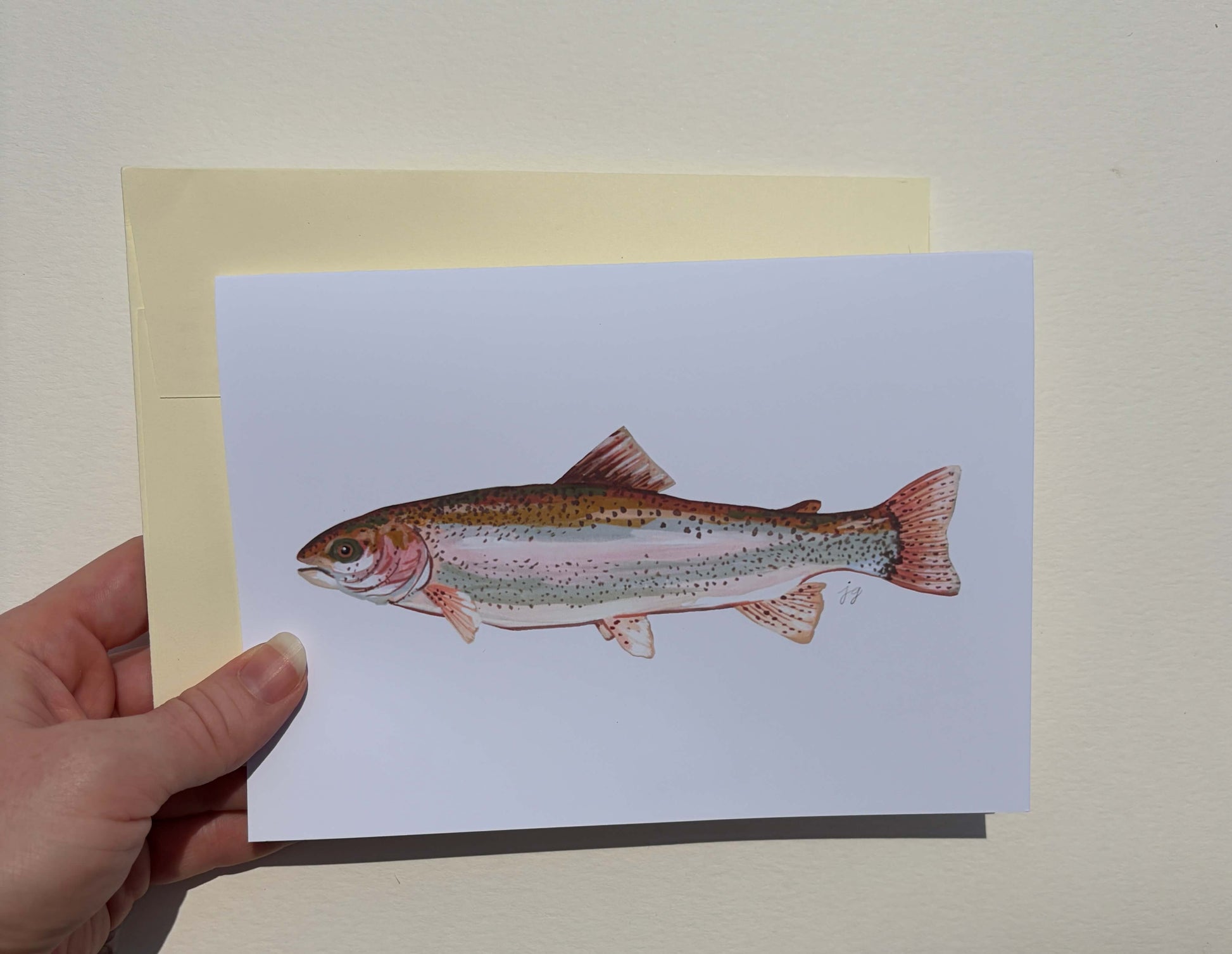 A hand holds a white art print or postcard featuring a detailed illustration of a rainbow trout, with a pale yellow backing card behind it against a light beige wall.