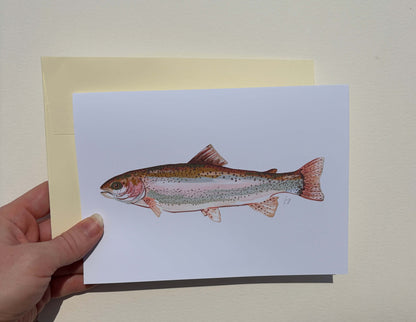 A hand holds a white art print or postcard featuring a detailed illustration of a rainbow trout, with a pale yellow backing card behind it against a light beige wall.