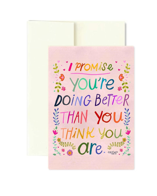 A pink greeting card with colorful hand-lettered text reading 'I promise you're doing better than you think you are' surrounded by small floral illustrations, shown with a white envelope behind it on a white background.
