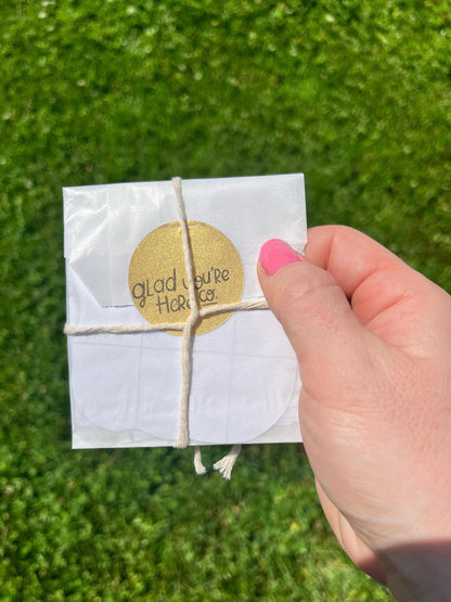 Hand holding a small white sticker package with a gold 'Glad You're Here' tag against a grass background