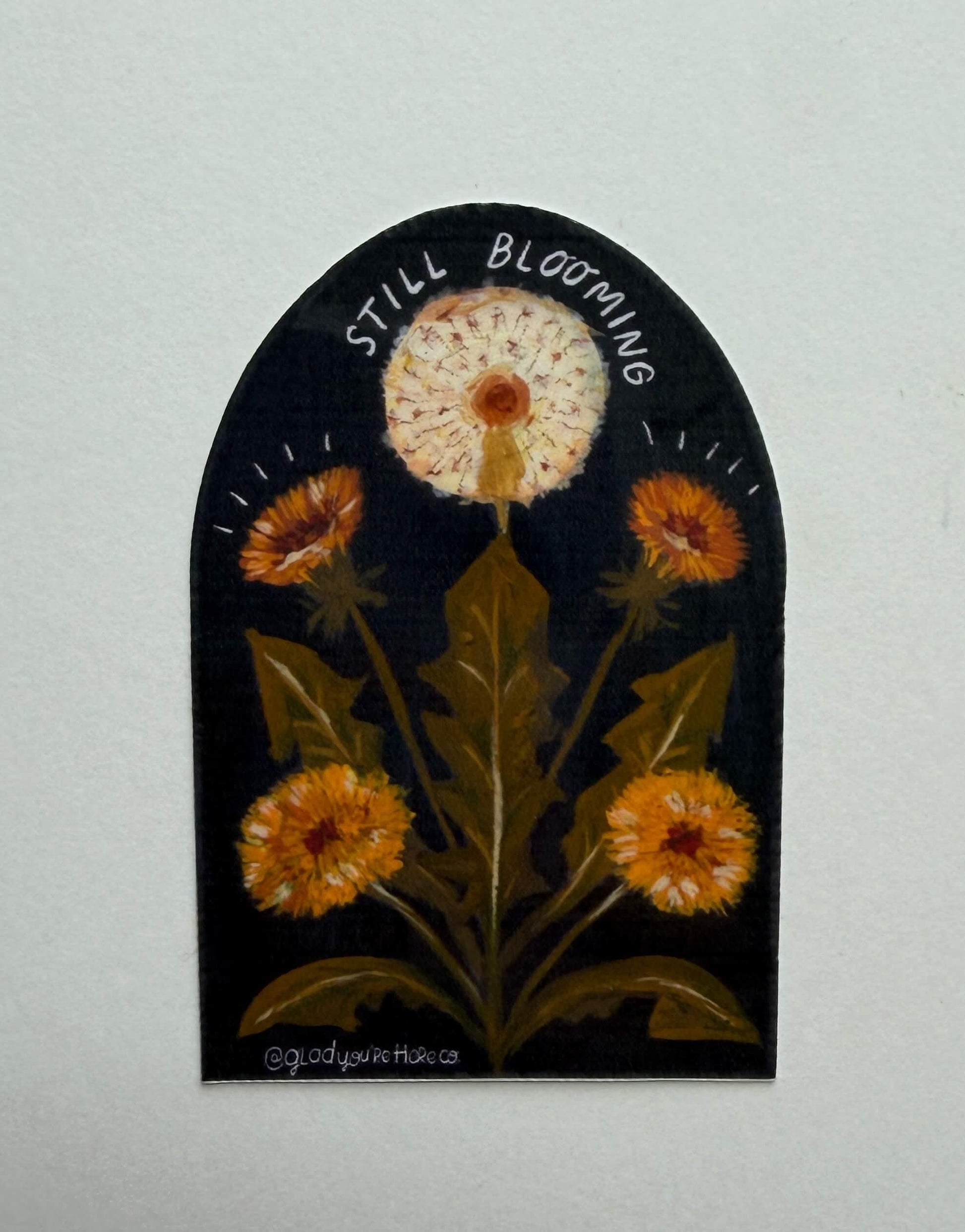 Decorative wall art with a dandelion and text 'Still Blooming' on a plain background