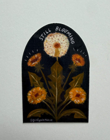 Decorative wall art with a dandelion and text 'Still Blooming' on a plain background