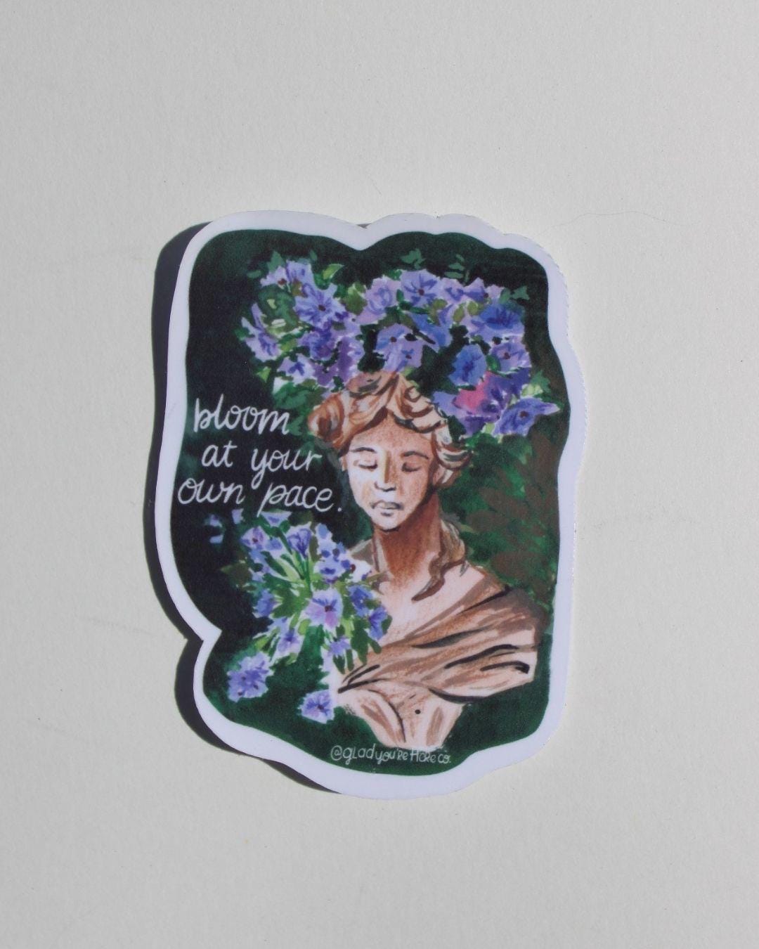Sticker with a woman's face surrounded by flowers and text 'bloom at your own pace' on a light gray background.
