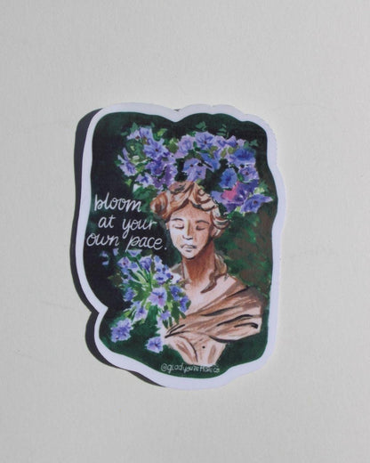 Sticker with a woman's face surrounded by flowers and text 'bloom at your own pace' on a light gray background.