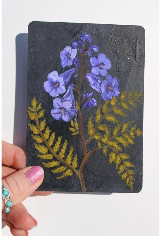 Hand holding a small painted card with purple flowers and green leaves on a white background