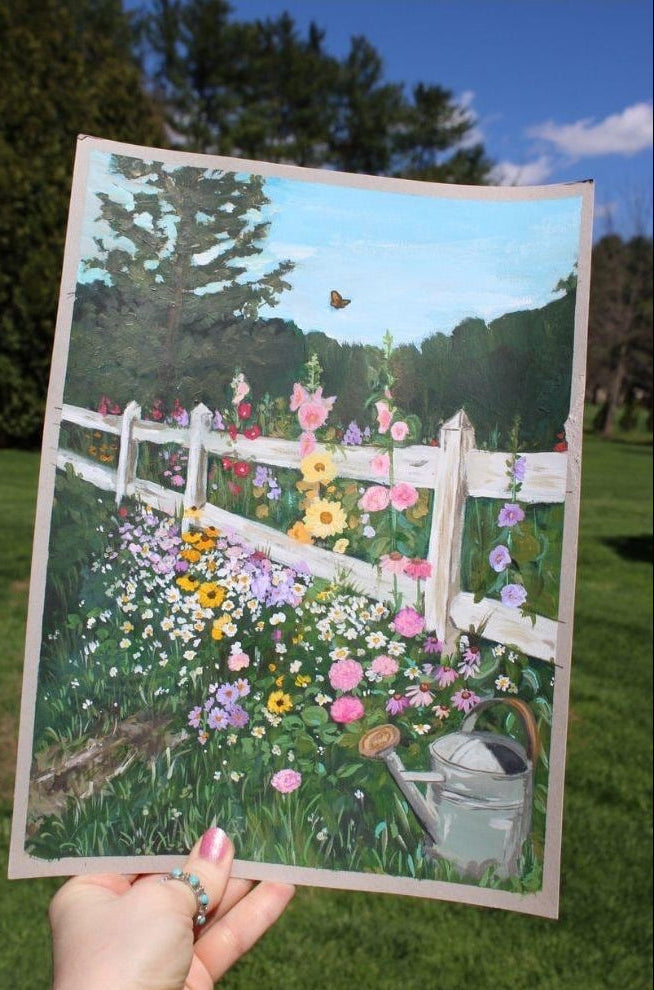 Hand holding a painting of a garden with flowers and a white fence.