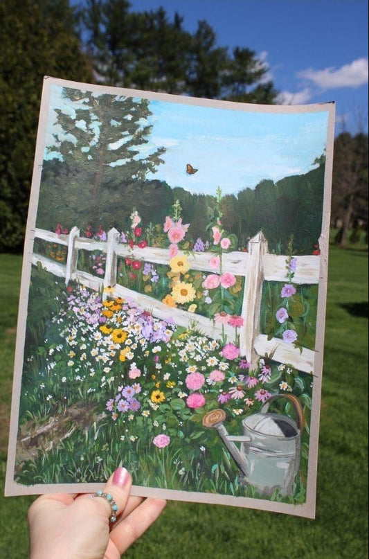Hand holding a painting of a garden with flowers and a white fence.