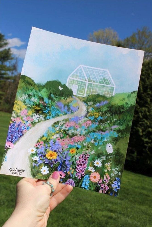 Hand holding an art print with a colorful illustration of a garden and greenhouse.