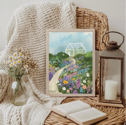 Framed artwork of a garden scene with flowers and a greenhouse, placed on a woven surface with a candle and lantern.