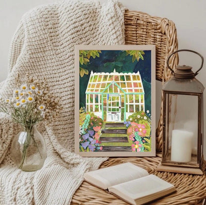 Framed collage print of a garden scene with flowers and a greenhouse, placed on a textured surface with a candle and lantern.