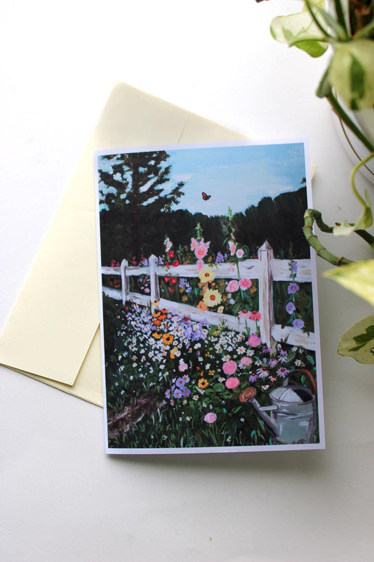 Greeting card with a garden scene featuring flowers and a white picket fence, placed on a white surface.
