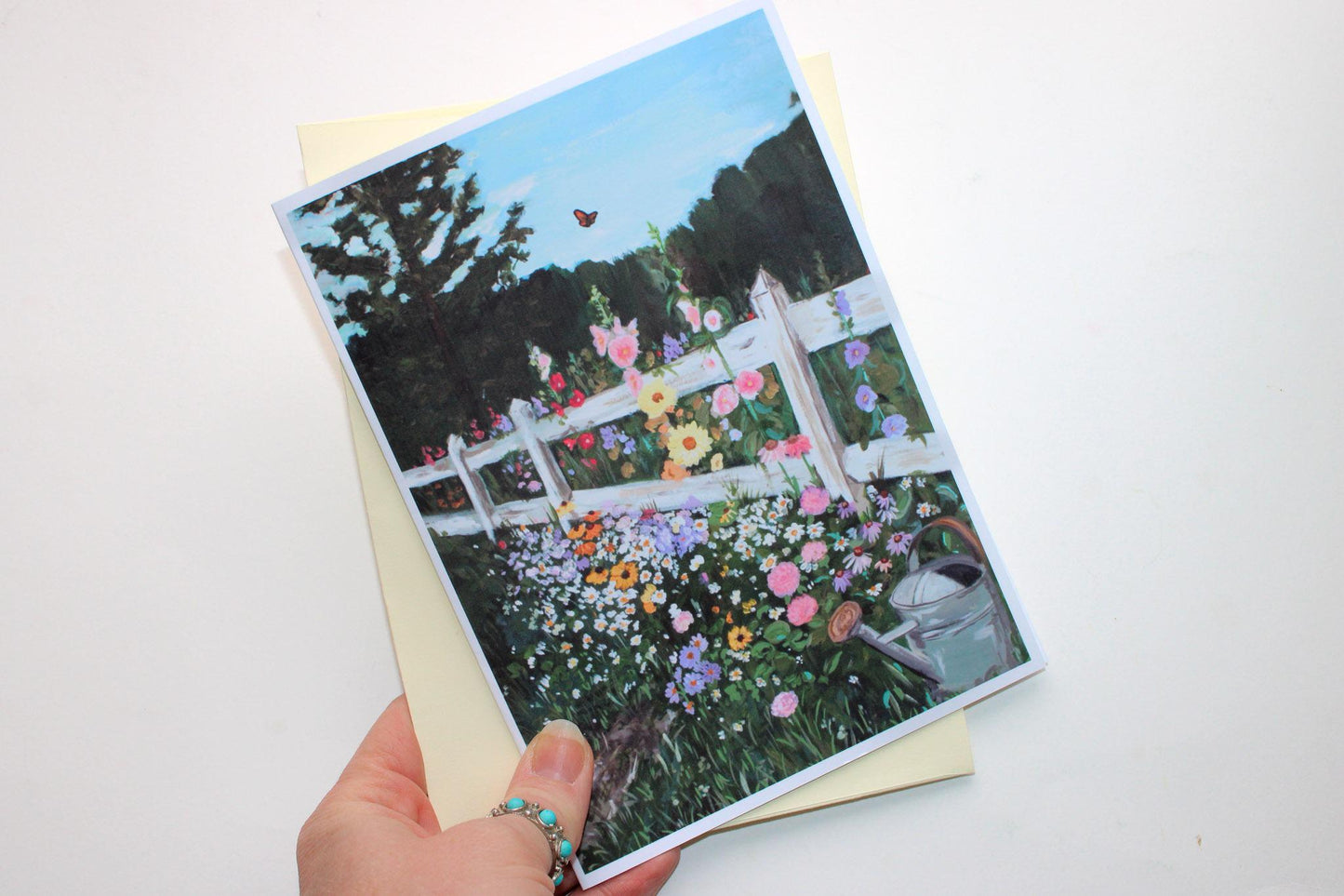 Greeting card with a garden scene featuring flowers and a white fence held by a hand on a white background