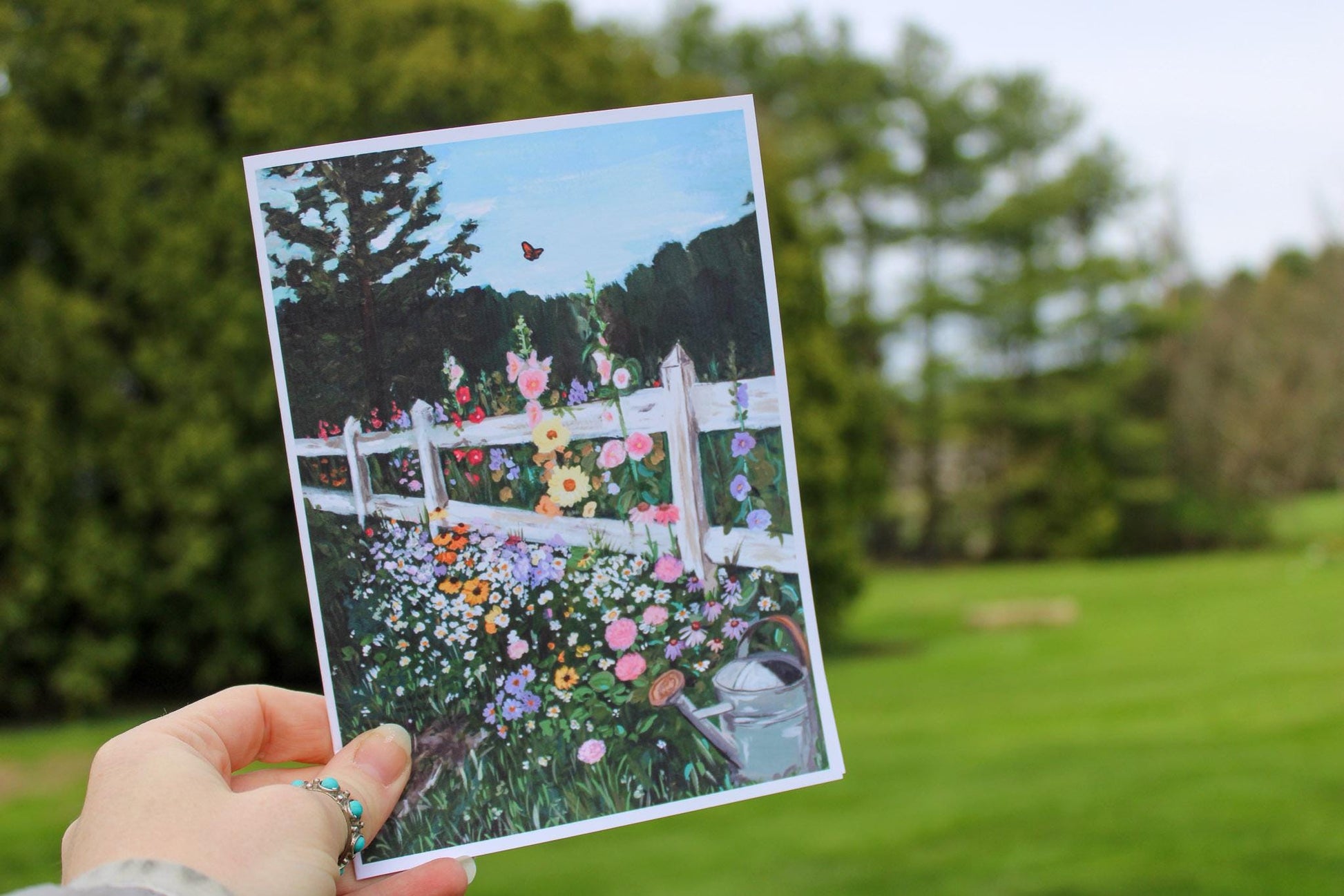 Hand holding a postcard with a garden scene featuring flowers and a white fence.