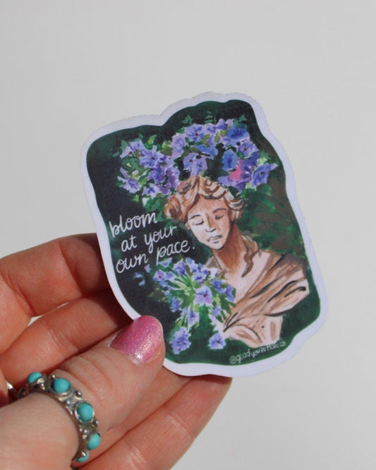 Sticker with a design of a woman with flowers and text held by a hand on a light gray background