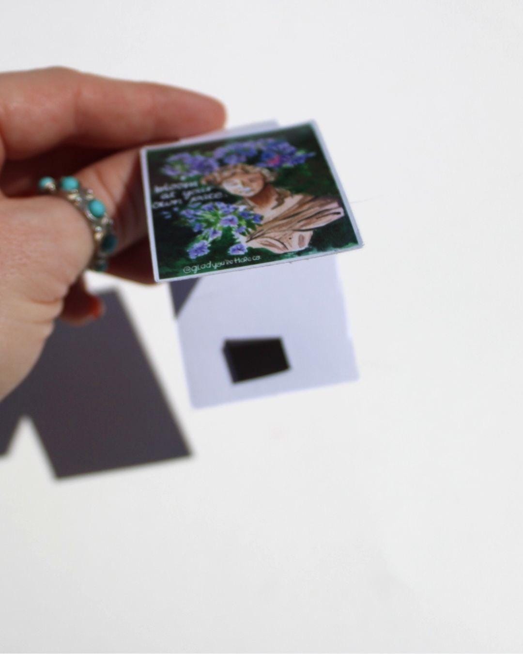 Hand holding a small magnetic bookmark with a garden statue and flowers on a white background