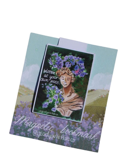Magnetic bookmark with a statue of woman's face wearing a floral crown and text 'Bloom at your own pace' on a scenic background.