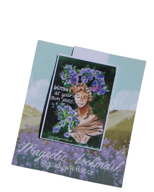 Magnetic bookmark with a statue of woman's face wearing a floral crown and text 'Bloom at your own pace' on a scenic background.