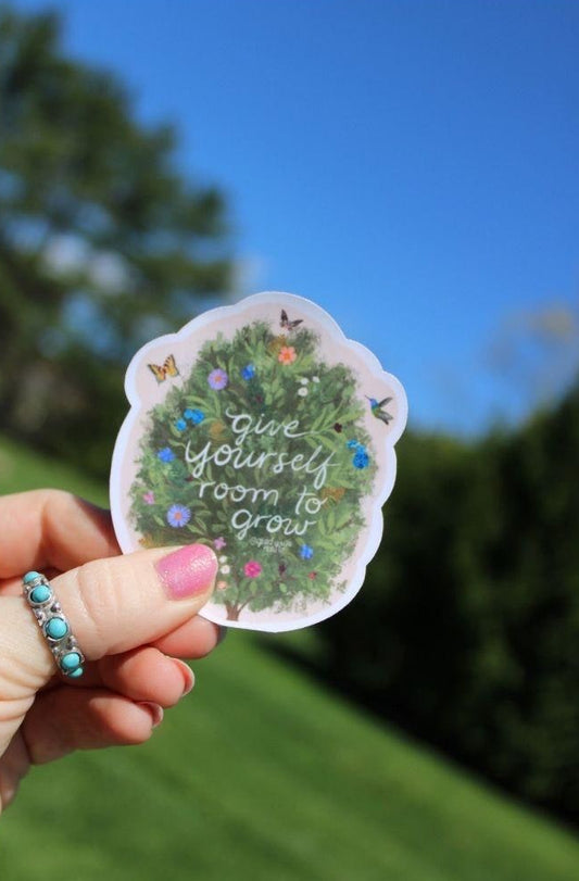 Hand holding a sticker with 'Give yourself room to grow' text against a blurred outdoor background