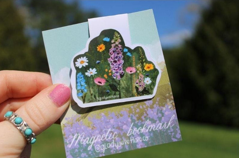 Hand holding a magnetic bookmark with floral design against a blurred natural background