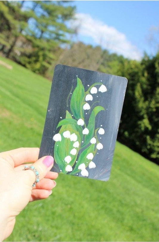 Hand holding a card with a painting of lily of the valley flowers against a natural background.