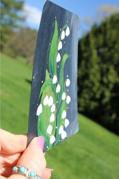 Hand holding a small painted canvas with a floral design outdoors.